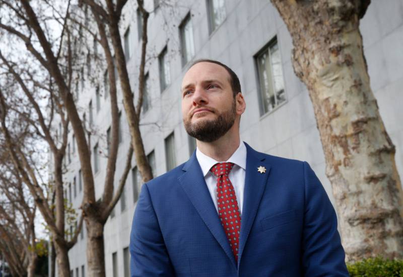 A young white man with a beard stands in a blue suit outside a gray building