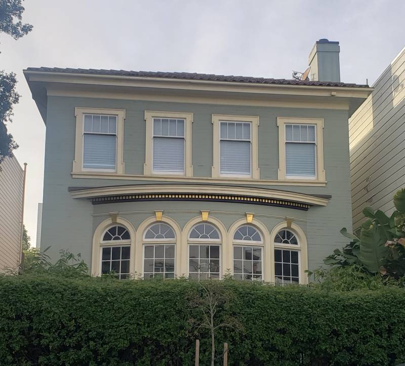 A two-story rectangular home with four identical windows on the top floors and a five curved windows protruding from the building on the lower level. Large neat hedges grow in front of the home.