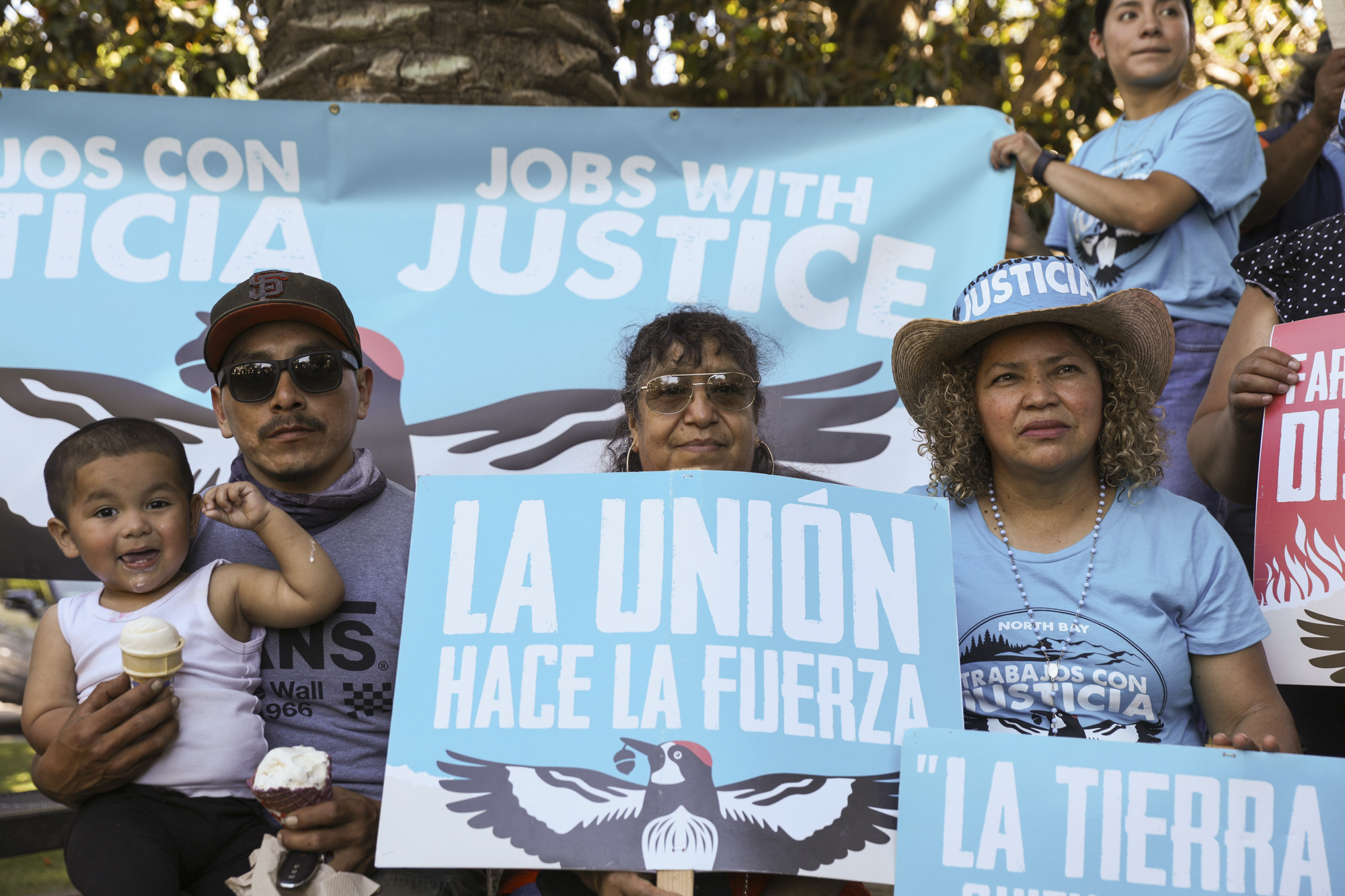 A group of people sit together holding signs reading "La Unión Hace La Fuerza" in an outdoor setting.