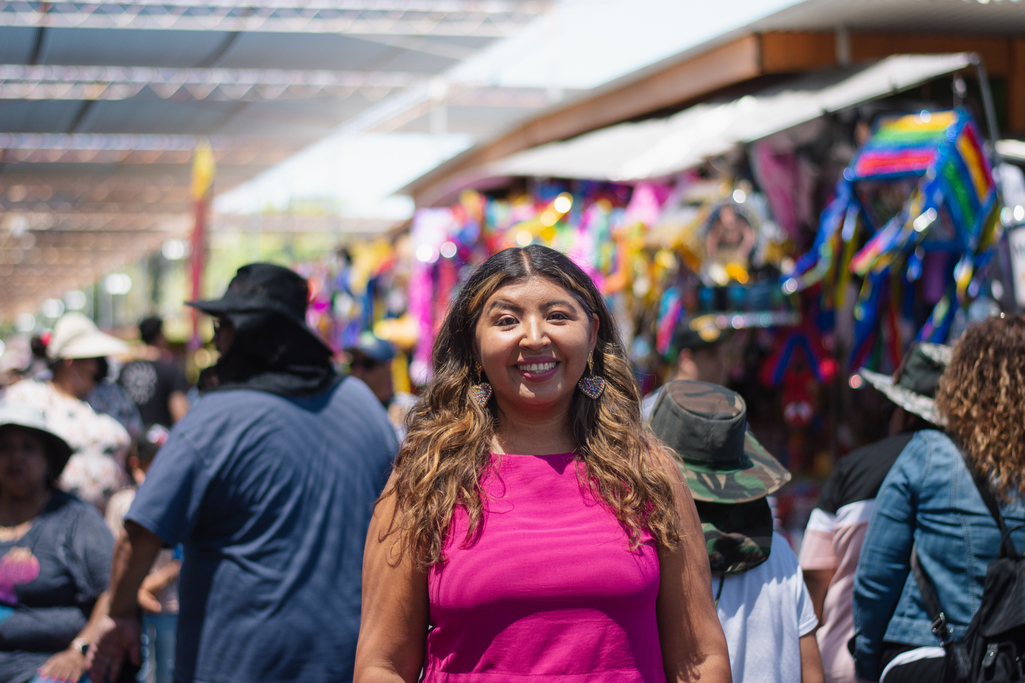 A person wearing earrings in a pink top smiles at the camera in a busy outdoor setting.