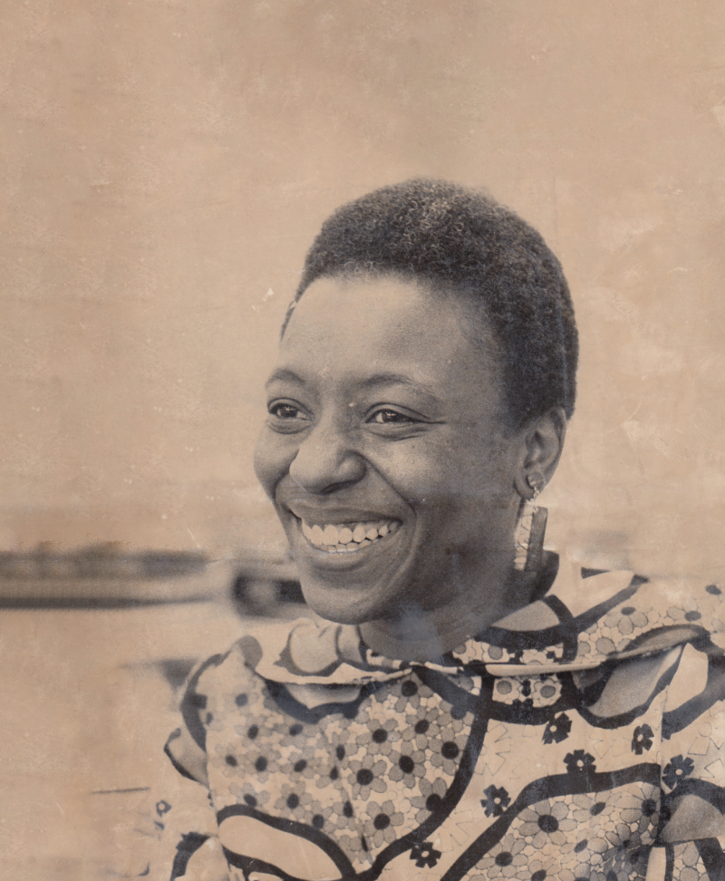 A Black woman with very short hair smiles broadly, looking off in the distance. She is wearing a 1970s-era blouse with lots of ruffles.